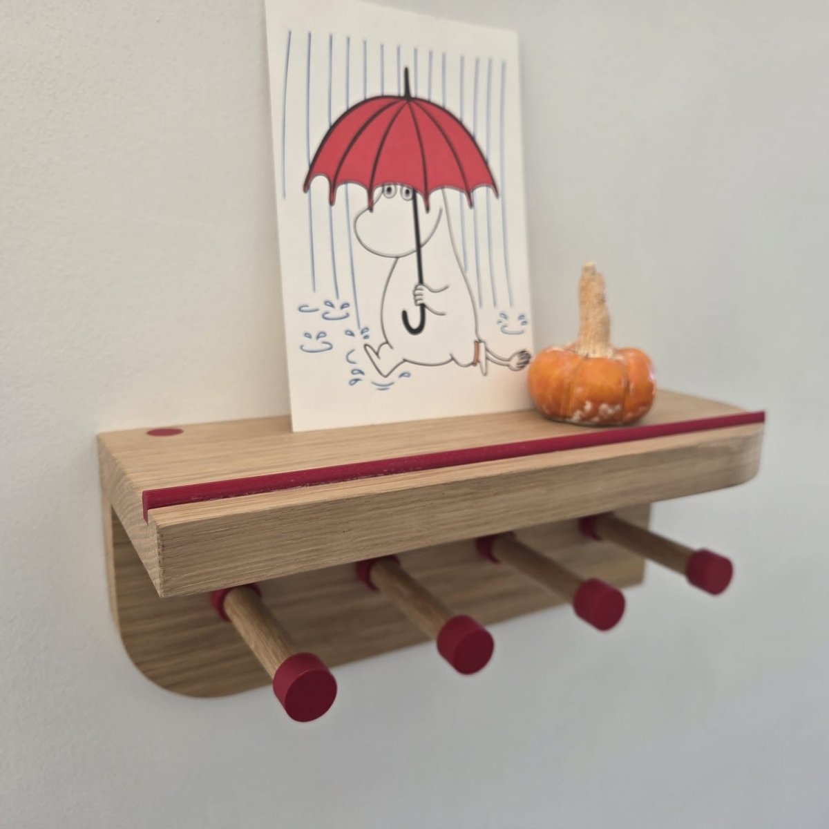  Oak wall-mounted key hook with four round wooden pegs tipped in red, and a slim red groove along the top shelf edge. The shelf holds a postcard of a cartoon moomin figure with a red umbrella and a small decorative ceramic pumpkin. 