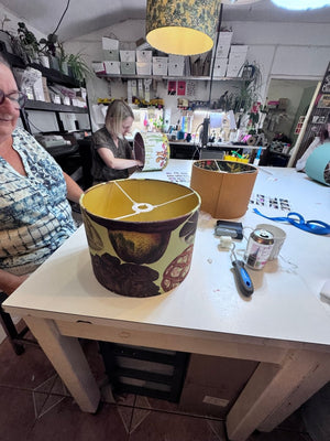 A person wearing glasses is seated at a white worktable in a cluttered craft or workshop space, while another person works in the background. The main subject in the foreground is a lampshade with a yellow interior and a brown floral pattern on the outside, alongside other lamp-making materials on the table.