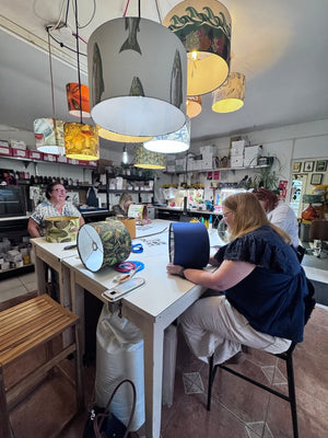 A woman in a navy blue top and beige pants sits at a white table in a craft workshop, making a double sided drum lampshade. Another woman is visible in the background near a lampshade.
