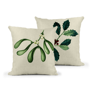 Pair of square linen-effect cushions featuring botanical Christmas designs. One cushion shows mistletoe with green leaves and white berries; the other displays holly with dark green spiky leaves and red berries, both on a neutral fabric background.
