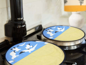 Two hob covers pictured on a cooker top. Circular design with seagulls flying on a bright blue background above yellow representing sand. .Oh Gully hob cover cooker Accessories Mustard and Gray Ltd Shropshire UK