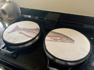Set of two. painted illustration of a salmon fish with green and pink colouring print on a beige linen circular hob cover with black hemming. Fish head on one hob pad and fish tail of the other hob pad. Pictured on a range cooker. Mustard and Gray Severn Salmon Circular Hob Cover